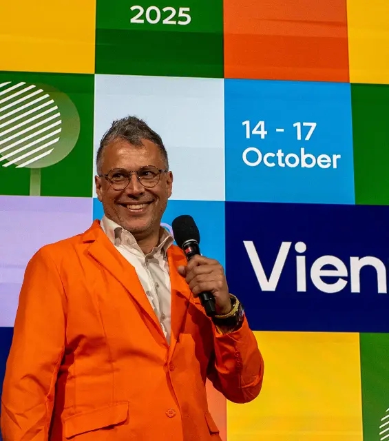 Speaker in an orange jacket holds a microphone on stage at DrupalCon Europe 2025, with the event branding for Vienna, 14–17 October, displayed behind him.