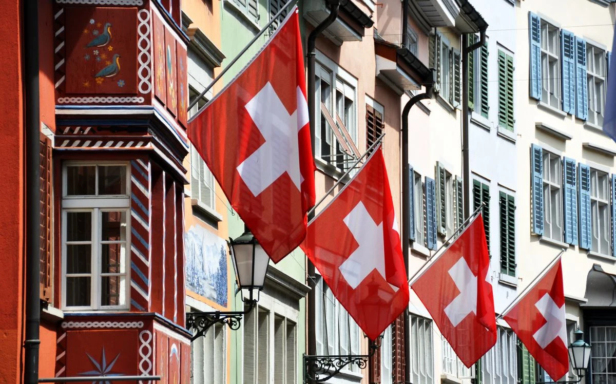 Colorful historic buildings line a street, each displaying red Swiss flags with white crosses, with decorative shutters, painted facades, and traditional architectural details visible in daylight.