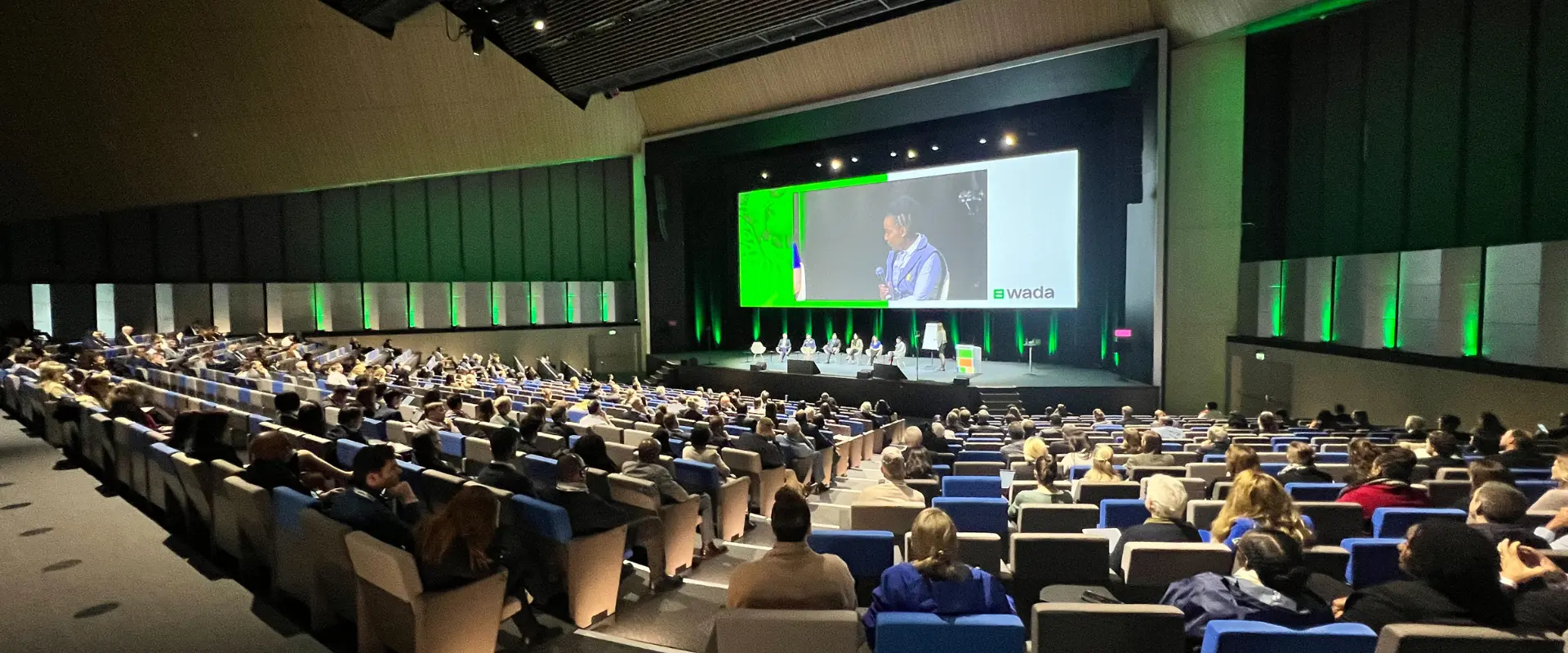 Conference delegates seated in a modern auditorium attending a symposium, with speakers on stage and a large presentation screen displaying a panel discussion.