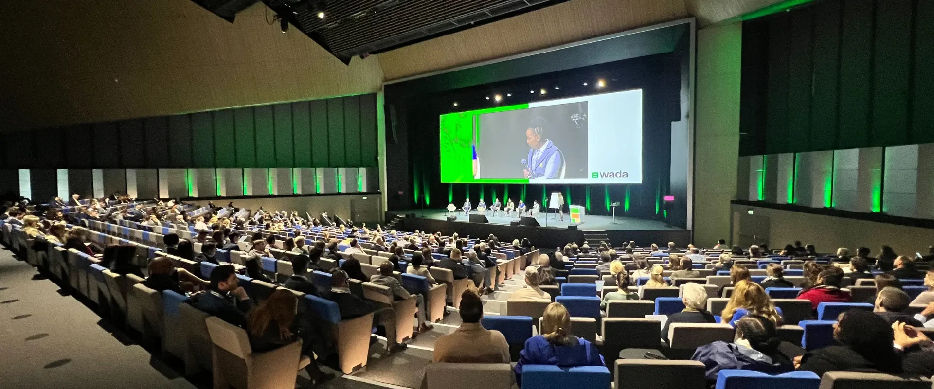 Conference delegates seated in a modern auditorium attending a symposium, with speakers on stage and a large presentation screen displaying a panel discussion.