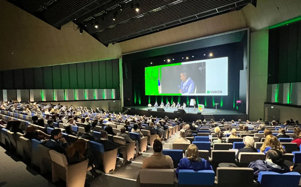 Conference delegates seated in a modern auditorium attending a symposium, with speakers on stage and a large presentation screen displaying a panel discussion.