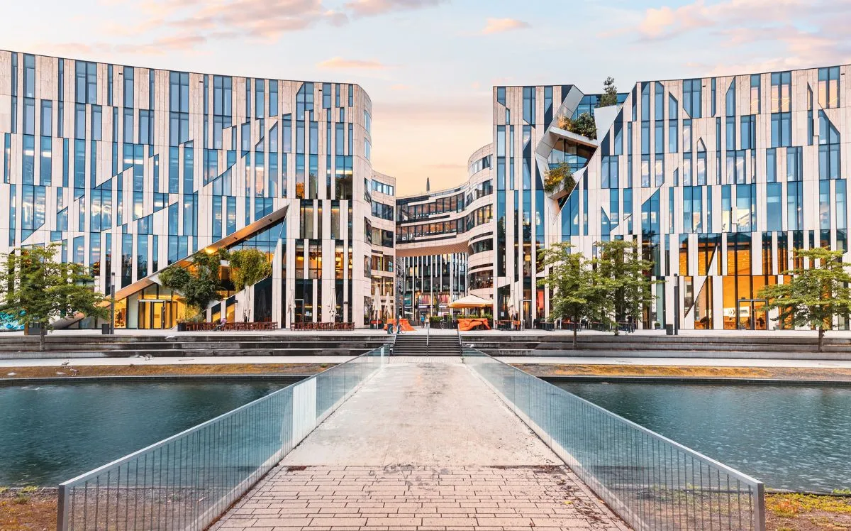 Modern office and business district in Düsseldorf featuring contemporary glass buildings, geometric architecture, landscaped water features, and a pedestrian bridge leading into the corporate complex at sunset.