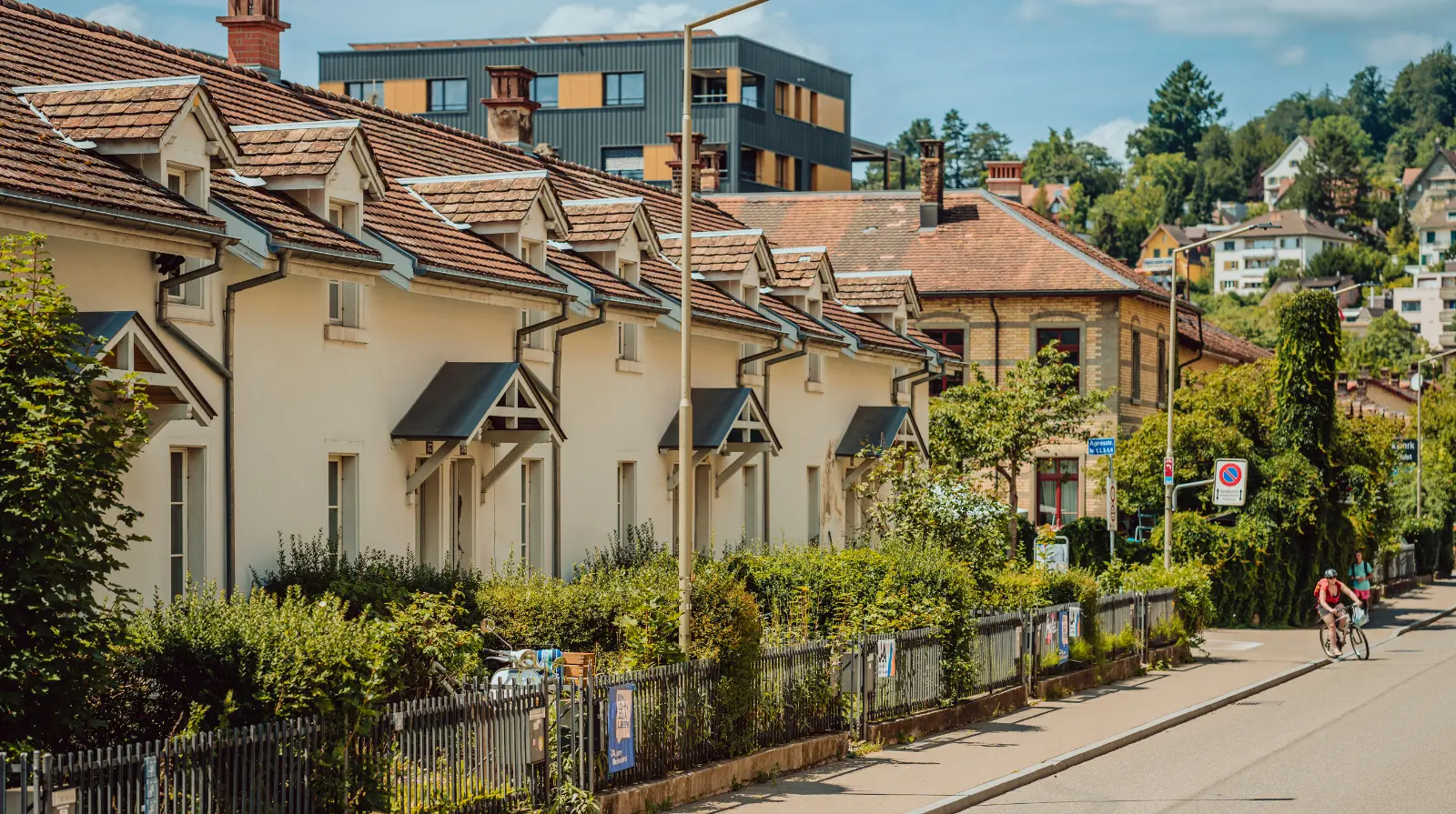 Quiet residential street in Winterthur lined with traditional Swiss houses, mature gardens and green hedgerows on a sunny day.