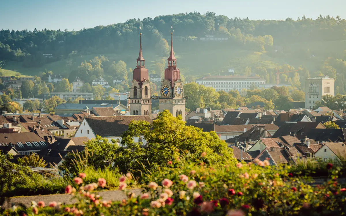 alt text for this image please16:07Panoramic view of Winterthur city centre featuring the twin towers of the Stadtkirche rising above rooftops, surrounded by lush green trees and rose gardens with wooded hills in the background.