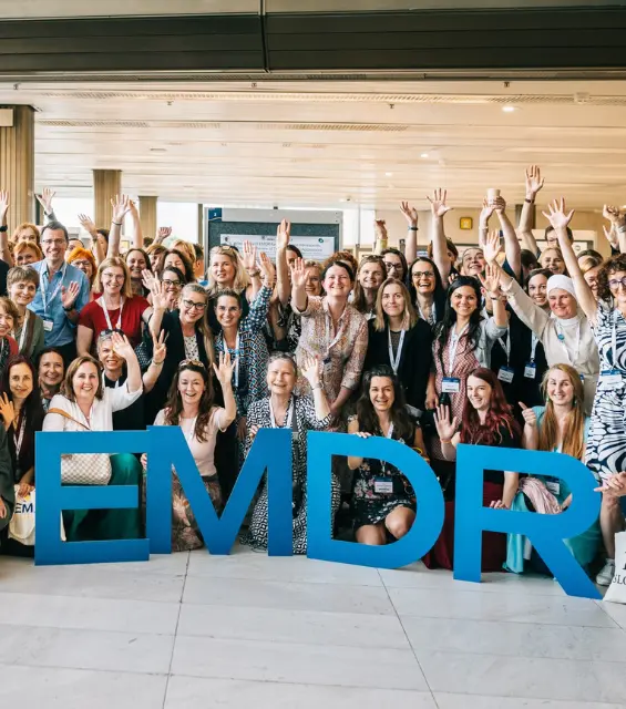 Group photo of approximately 100 conference attendees posing together in a bright, modern venue, many waving and raising their hands, with large blue 3D letters spelling 