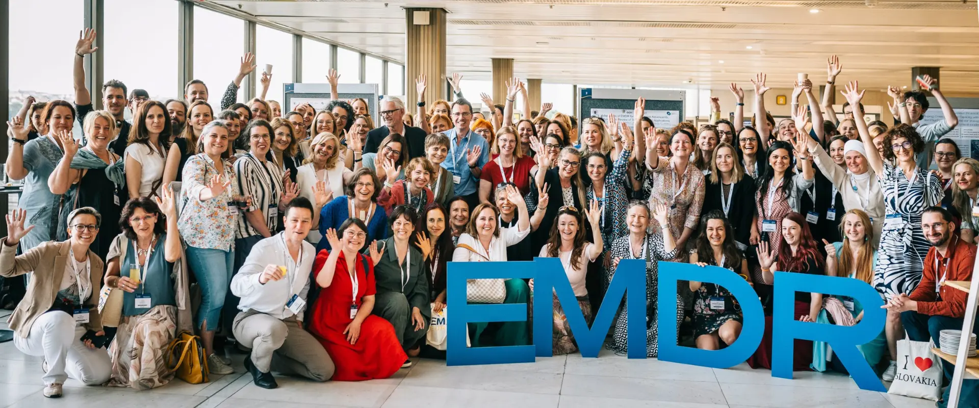 Group photo of approximately 100 conference attendees posing together in a bright, modern venue, many waving and raising their hands, with large blue 3D letters spelling 