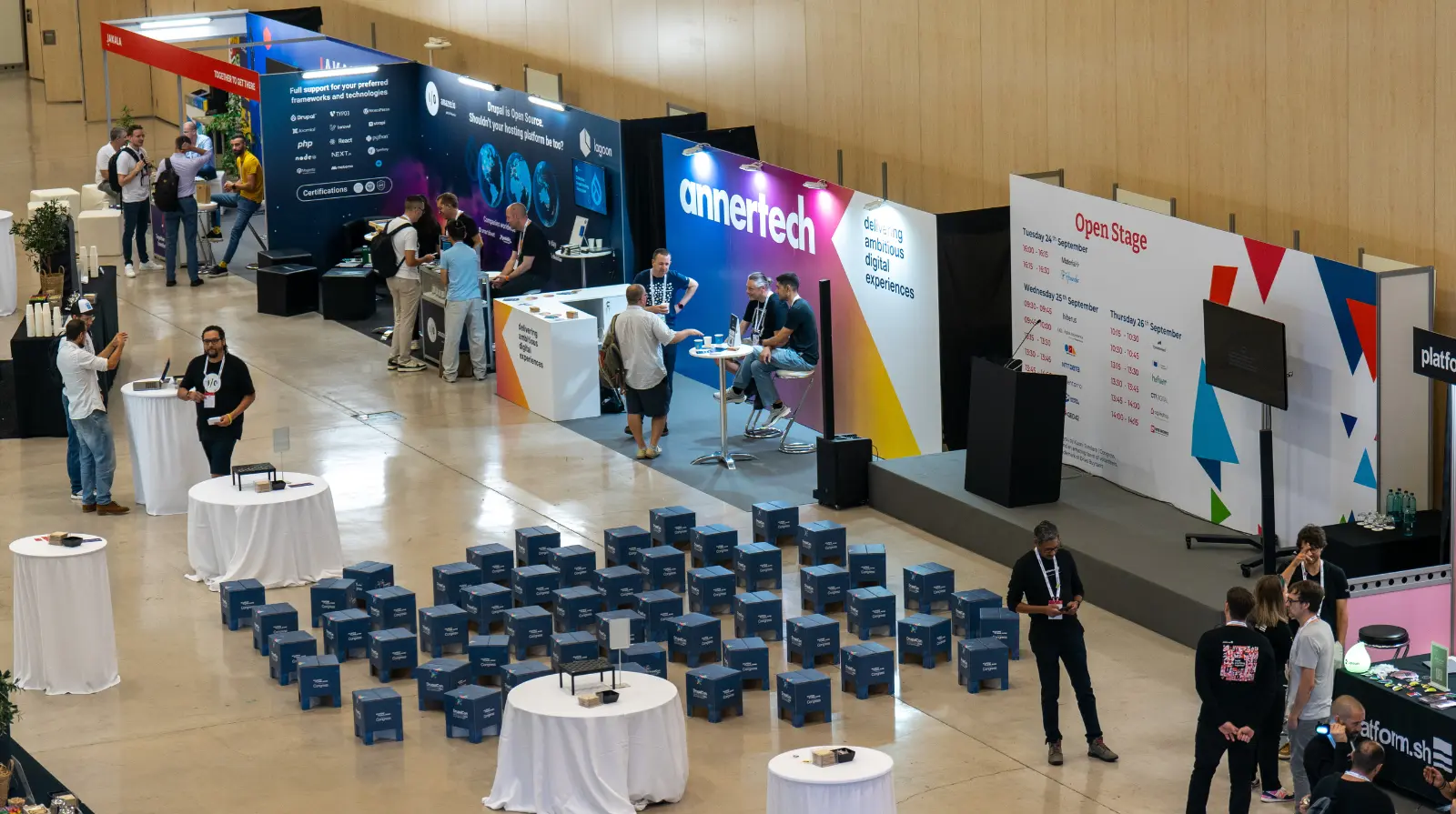 Overhead view of the DrupalCon Europe exhibition hall, showing sponsor stands, the Open Stage schedule board, and seating made from branded cardboard stools.