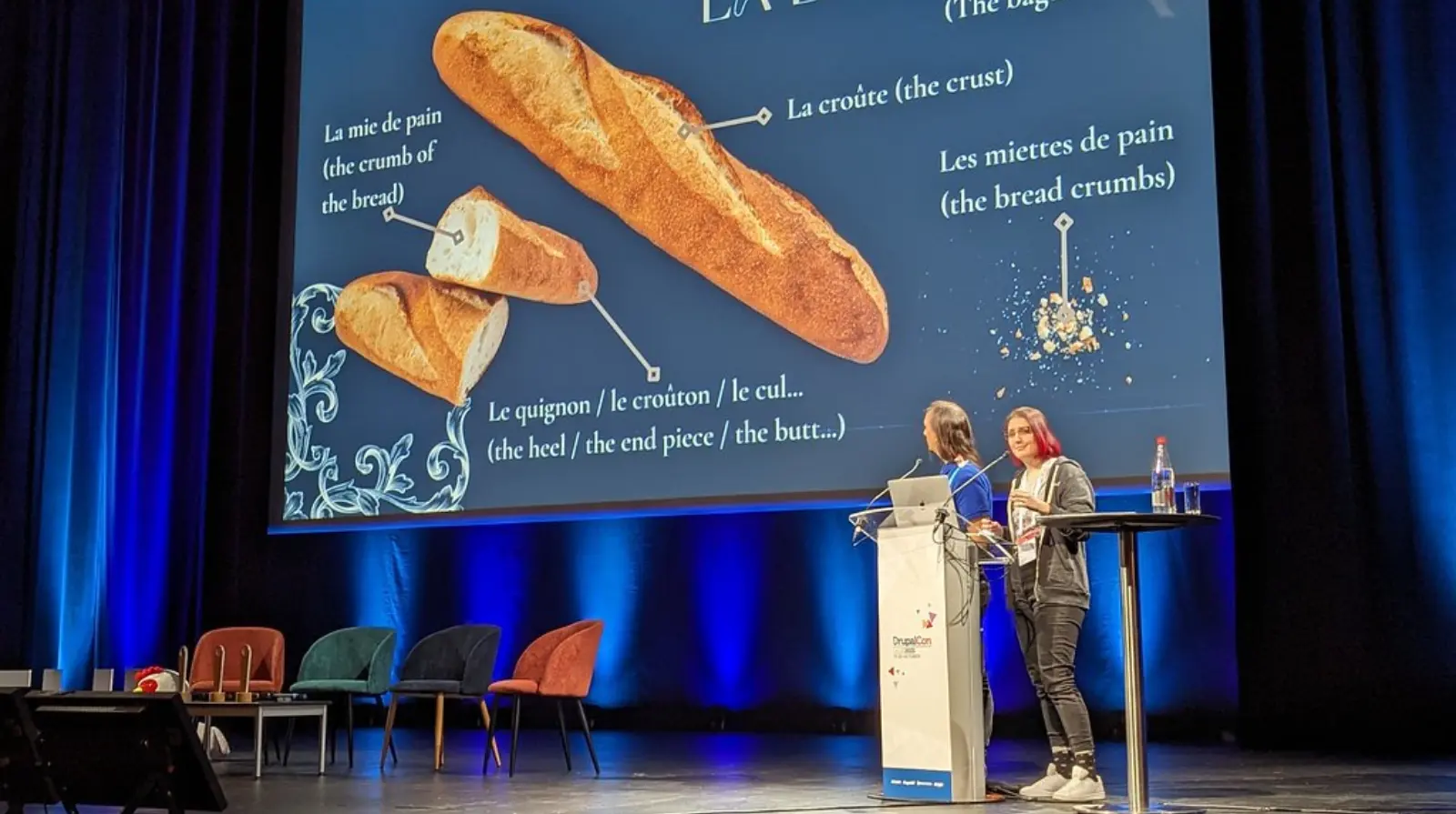 wo speakers present on the DrupalCon Europe Lille 2023 main stage, with a slide explaining the parts of a French baguette displayed behind them.