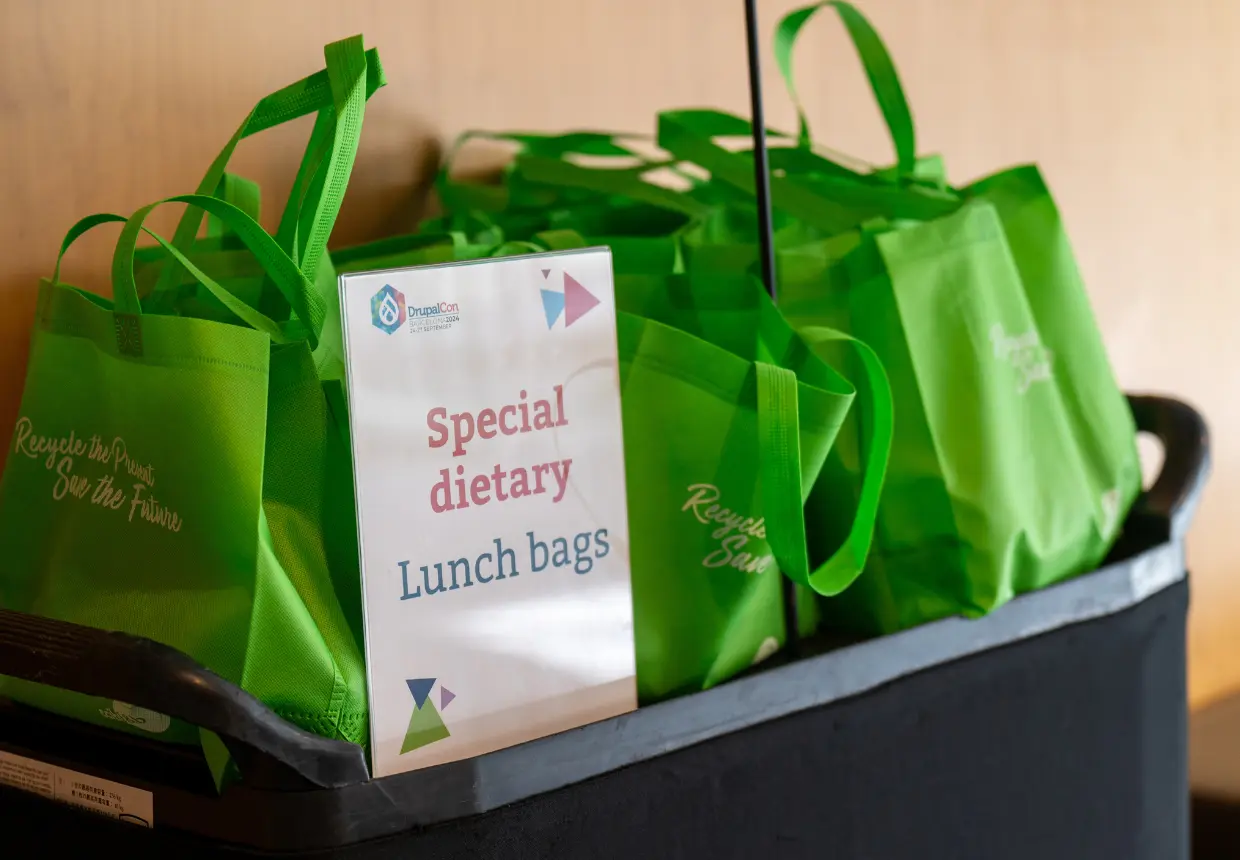 Reusable green DrupalCon Europe Barcelona 2024 lunch bags prepared for delegates with special dietary requirements, printed with the slogan 