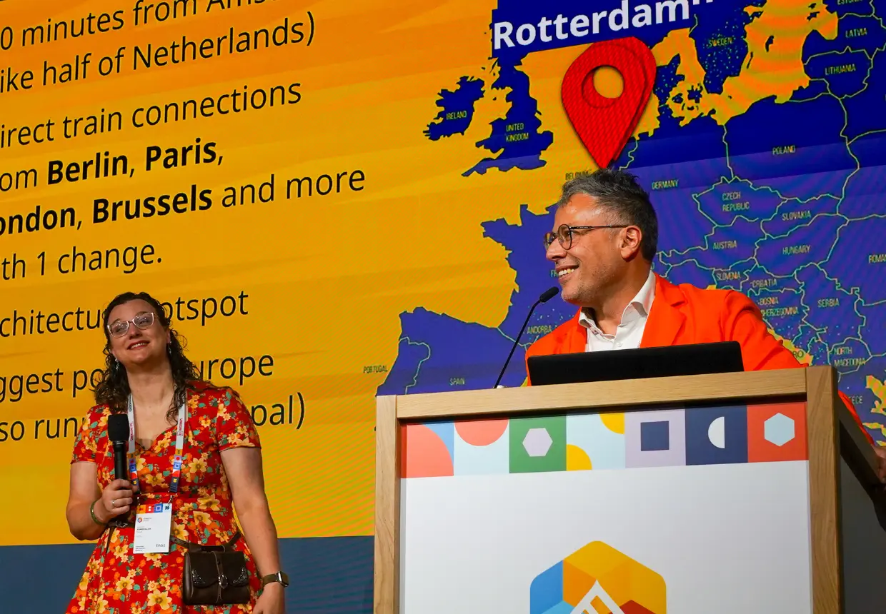 Speakers on the DrupalCon Europe stage present Rotterdam 2026, highlighting direct rail connections from Berlin, Paris, London and Brussels as low-emission travel options for delegates.