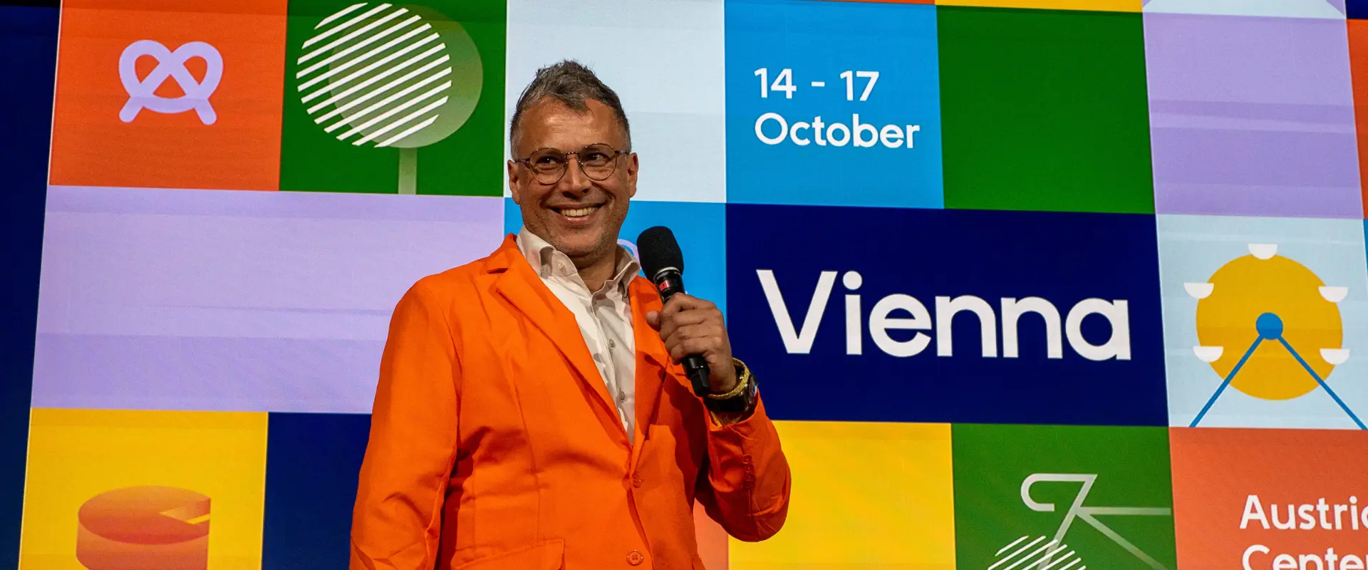 Speaker in an orange jacket holds a microphone on stage at DrupalCon Europe 2025, with the event branding for Vienna, 14–17 October, displayed behind him.