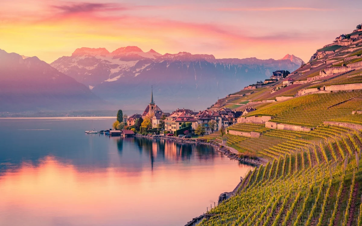Terraced vineyards sloping to Lake Geneva at sunset, with a lakeside village church and snow-capped Alps