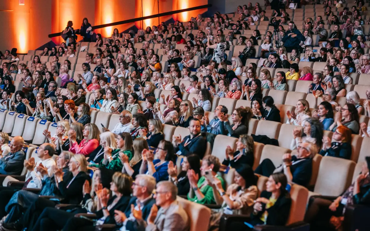 Large conference audience applauding from tiered seats in an auditorium, many wearing lanyards, with orange-lit walls in the background.