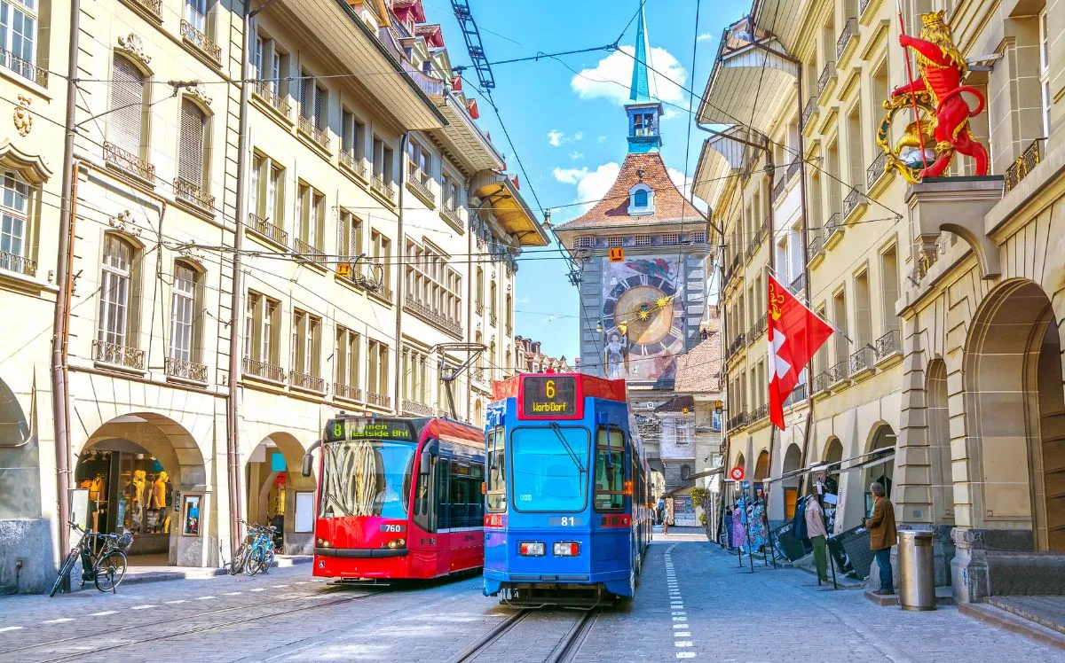 Historic street in Bern, Switzerland, with colorful trams running along cobblestone tracks, traditional buildings with arcades, Swiss flags, and the Zytglogge clock tower in the background under a clear blue sky.