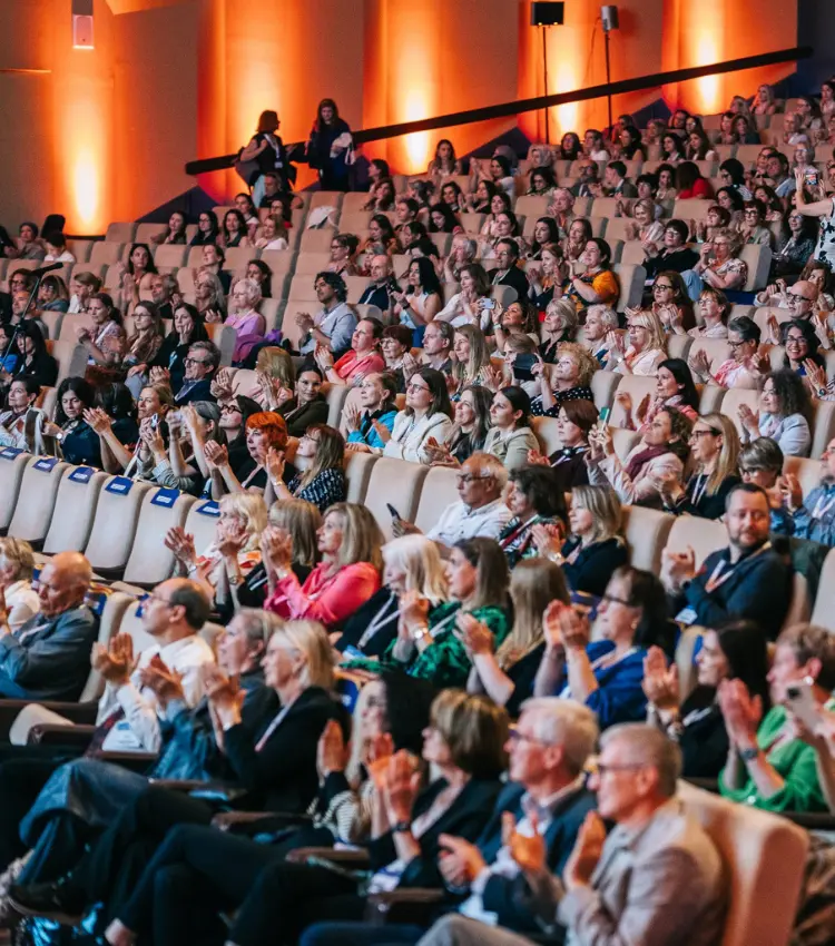 Large conference audience applauding from tiered seats in an auditorium, many wearing lanyards, with orange-lit walls in the background.