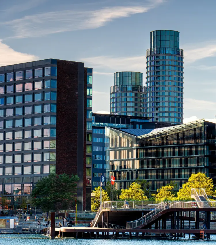 Modern glass office and hotel buildings line the Copenhagen waterfront, with a harbour pier in the foreground and the Danish flag visible among autumn trees, reflecting the city