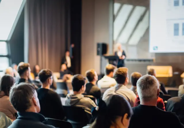 Delegates seated in a conference hall watching two speakers present on stage
