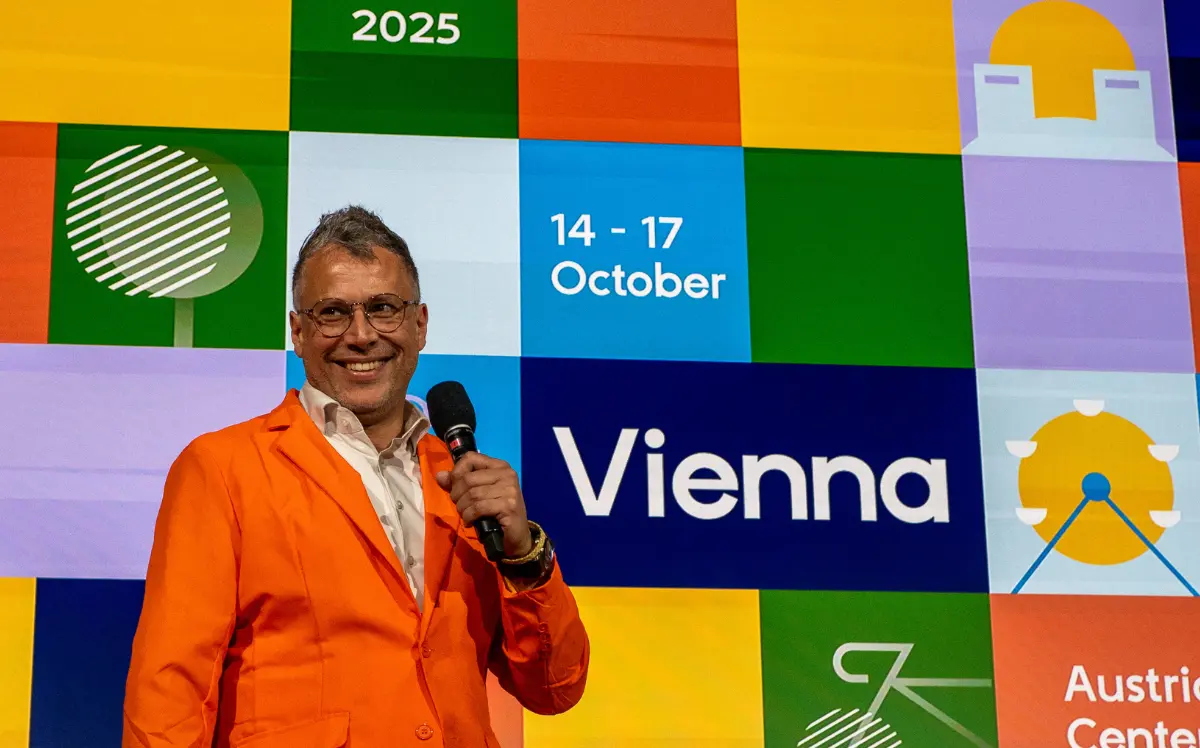 Speaker in an orange jacket holds a microphone on stage at DrupalCon Europe 2025, with the event branding for Vienna, 14–17 October, displayed behind him.
